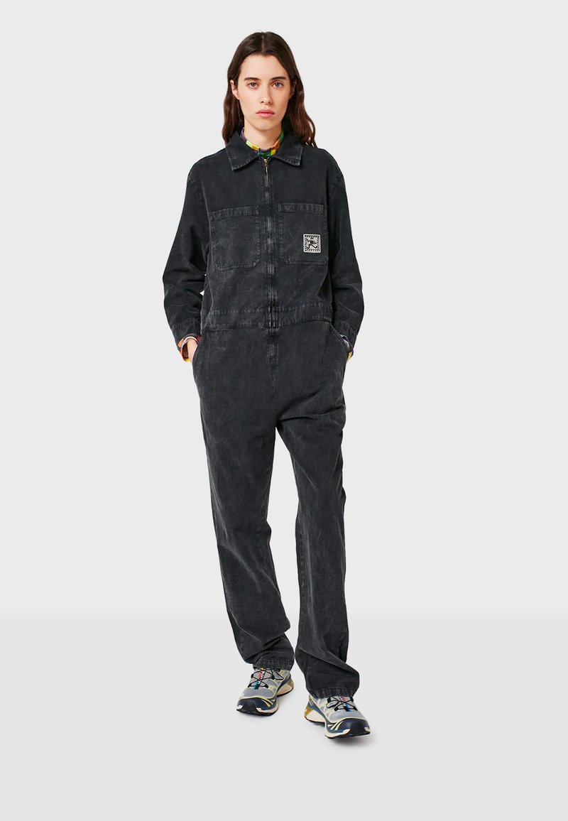 Grahamfarbener Jeans-Overall mit Reißverschluss vorne, langen Ärmeln und einer Brusttasche mit einem geometrischen Logoschriftzug. Kombiniert mit mehrfarbigen Sportschuhen.