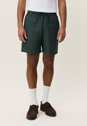 Uomo che indossa pantaloncini verdi scuro con cordoncino, calzini bianchi e scarpe stringate in camoscio marrone, in piedi davanti a uno sfondo neutro.