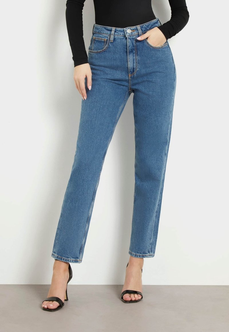 Femme portant un jean bleu taille haute à jambes droites, un haut noir à manches longues et des talons hauts noirs ouverts à l'avant, debout sur fond blanc.