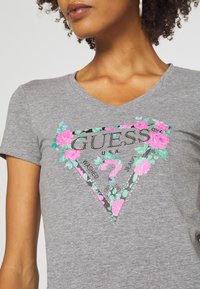 Kvinde iført en grå V-hals T-shirt med et blomsteragtigt pink og grønt Guess-logo med et spørgsmålstegn foran.