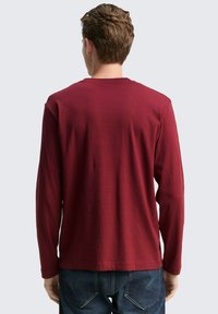 Man met kort haar, gekleed in een effen bordeauxrode langemouwen shirt en blauwe spijkerbroek, staand met zijn rug naar de camera tegen een lichte achtergrond.