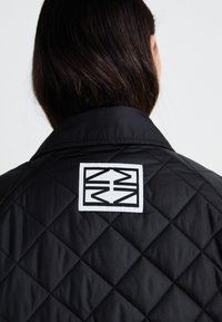 Veste matelassée noire avec un grand patch carré blanc présentant un logo géométrique noir au dos. Texture lisse, col ajusté.
