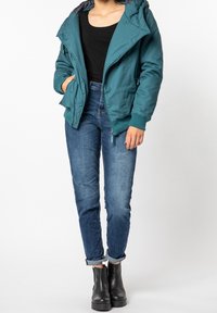 Veste zippée teal avec capuche et poignets élastiques, portée sur un haut noir et un jeans bleu, accompagnée de bottines noires.