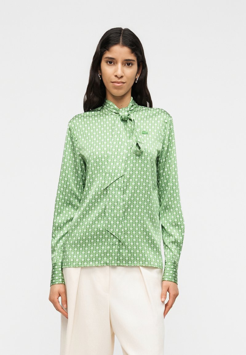 Femme portant un chemisier vert à motifs à manches longues avec un nœud noué au col et un pantalon ample de couleur crème, debout devant un fond uni.