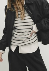 Schwarze Bomberjacke über einem gestreiften, kuscheligen Pullover mit schwarz-weißen Streifen, getragen mit einem weißen Hemd mit Kragen und schwarzen Hosen.