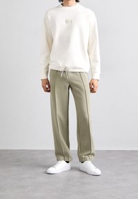 Sudadera de color beige claro con un pequeño logo, combinada con pantalones de chándal verde oliva que cuentan con puños elásticos y cintura ajustable con cordón. Zapatillas blancas.