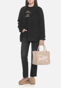 Cappotto di lana nero con due bottoni, maglione nero con scritte dorate, denim scuro, borsa di velluto a coste beige con scritte bianche e scarpe slip-on color cammello.