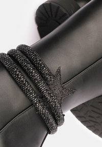 Botín de cuero negro hasta el tobillo que presenta una correa negra texturizada y brillante con un acento en forma de estrella. La suela es rugosa y está diseñada para ofrecer agarre.