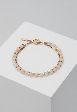 Bracelet doré avec de petites perles blanches facettées et un fermoir homard, présenté sur un piédestal géométrique blanc sur un fond gris.