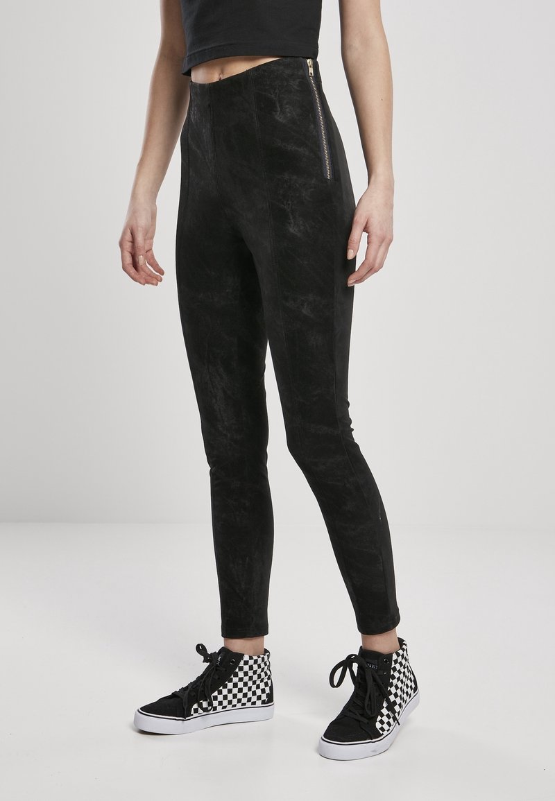 Partie inférieure du corps portant des leggings en velours noirs avec une fermeture éclair latérale et des baskets montantes à carreaux noir et blanc sur un fond gris de studio.