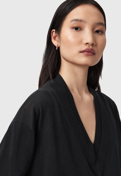 Jeune femme aux longs cheveux foncés portant un pull noir à col en V et une petite boucle d'oreille, expression neutre, sur un fond gris clair.