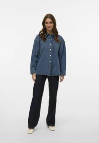 Denim shirt in dark blue, button-down front, two chest pockets, long sleeves, paired with dark blue wide-leg jeans and light shoes.