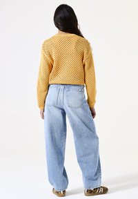 Un pull en tricot jaune avec un motif en tricot ouvert, associé à un jean large bleu clair et des chaussures à motif, vu de dos.