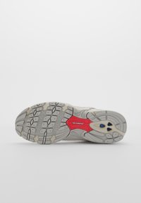 Semelle d'une basket New Balance grise et beige avec une accroche texturée, un patch logo rouge et des accents circulaires bleus sur un fond uni.