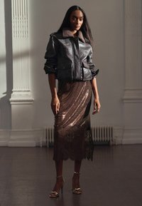 Frau trägt eine dunkle Lederjacke mit Pelzkragen, einen schimmernden, mit Spitze verzierten Midirock und metallische High-Heels und steht drinnen.