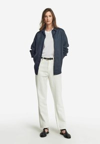 Marinblå linne skjorta över en vit t-shirt, i kombination med vita raka byxor och svarta flätade sandaler. Enkel, modern design.