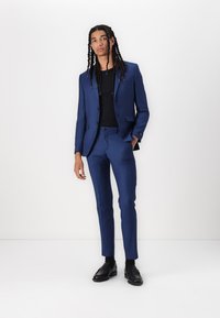 Costume bleu marine avec design ajusté, veste à un bouton, pantalon à taille plate et chaussures noires, associé à un haut sombre.