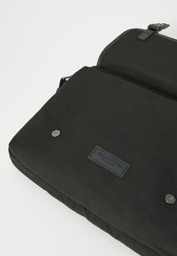 Borsa per laptop nera con una texture in tela opaca, dotata di una patch con logo e chiusura a bottone metallico. Design compatto con tracolla regolabile.