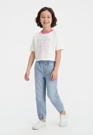 Kind met kort krullend haar dat glimlacht, draagt een wit cropped T-shirt met print, lichtblauwe spijkerbroek met elastische taille en witte instappers, staat rechtop.