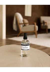 Bottiglia di vetro trasparente contenente un liquido con un tappo a spruzzo nero. L'etichetta riporta "N°09 Lemon Notes." Sistemata su un tavolo di colore chiaro e texture evidente.