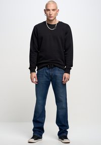 Felpa nera in cotone, jeans in denim blu e sneakers nere. Gli accessori includono una collana di catena d'argento e anelli.