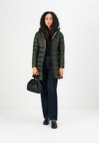 Olijfgroene pufferjas met een capuchon, ritsclosure en horizontale quilt; gecombineerd met donkerblauwe flared jeans en een kleine zwarte handtas.