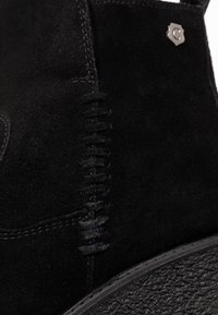 Svart mocka ankelstövel med en texturerad gummisula, vertikala sömdetaljer och en metallisk accent nära toppen.