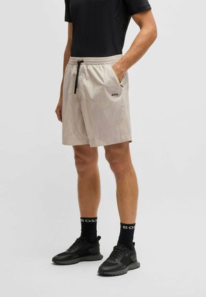 Homme portant un short beige avec le logo BOSS, des chaussettes noires, des baskets noires et une chemise noire, debout avec une main dans la poche.