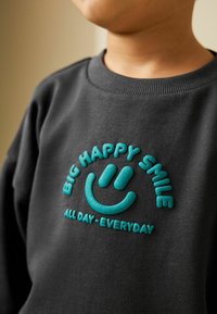 Tmavě šedá mikina s vyvýšeným akvamarínovým textem "BIG HAPPY SMILE" a grafikou smajlíka. Pohodlný design s kulatým výstřihem.