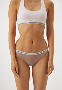 Calvin Klein könnyű pamut keverék bralette fehér színben, logós pánttal; világosbarna, átlátszó csipkéből készült bikinit visel.