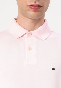 Lichtroze poloshirt met een klassieke kraag, twee knopen en een klein merkembleem in rood en blauw op de linkerborst. De textuur lijkt ademend.