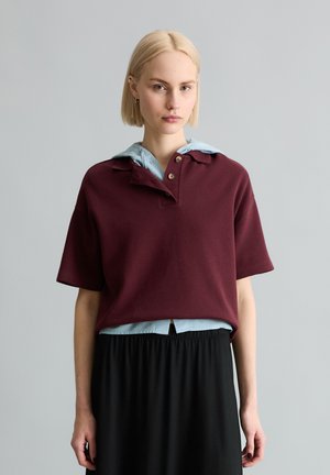 Blonde Frau trägt ein kurzärmeliges, kastanienbraunes Poloshirt über einem hellblauen Kapuzenpullover und einem schwarzen Rock, steht vor einem grauen Hintergrund.