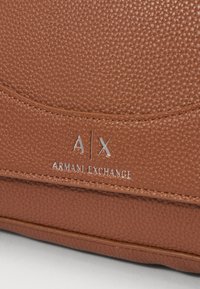 Brun teksturert lærveske med et metallisk A|X-logo og præget "ARMANI EXCHANGE"-detalj, med et elegant, minimalistisk design.