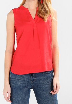 Mujer con una blusa roja sin mangas con escote en V y vaqueros azules contra un fondo claro y liso.