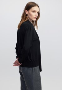 Zwarte gebreide cardigan met een ontspannen pasvorm, open voorkant en geribbelde zoom. Model draagt grijze, textuurvolle broek. Eenvoudig en strak ontwerp.