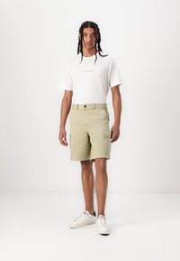 Light green cargo shorts with side pockets, paired with a white T-shirt featuring black text. Model wears white sneakers.