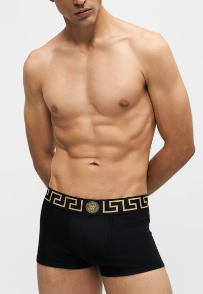 Torse masculin portant un boxer noir avec une ceinture à motif doré et un logo tête de Méduse au centre avant.
