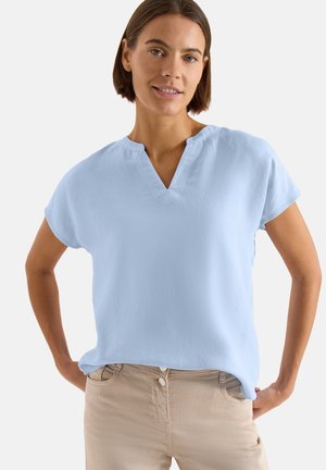 Femme aux cheveux bruns courts porte un chemisier bleu clair à manches courtes et un pantalon beige, debout mains sur les hanches devant un fond uni.