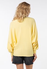 Gelber übergroßer Pullover mit Ballonärmeln, gerippten Bündchen und strukturiertem Stoff, kombiniert mit dunklen Denim-Shorts. Rückansicht gezeigt.