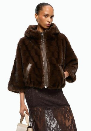 Mujer con chaqueta de piel marrón oscuro y falda negra de encaje, sosteniendo un bolso beige, de pie frente a un fondo blanco liso.