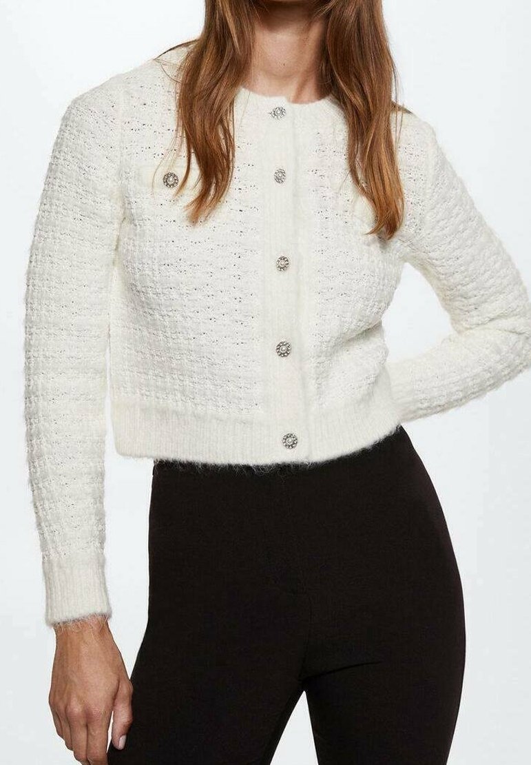 Frau in einem strukturierten weißen verkürzten Cardigan mit dekorativen Knöpfen und schwarzen, eng anliegenden Hosen vor einem schlichten Hintergrund.