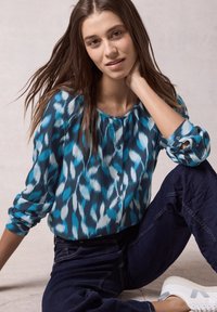 Camicia blu con motivo a fantasia, con maniche lunghe e scollatura rotonda. Indossata con jeans scuri e sneakers bianche, seduta contro uno sfondo neutro.