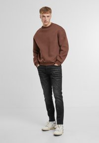 Sudadera marrón con hombros caídos, combinada con jeans negros desgastados que presentan manchas de color claro. Zapatillas blancas completan el look.