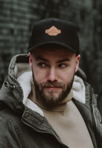 Fekete trucker sapka barna bőrrel ellátott "BLACKSKIES" logóval. Zöld kabáttal és bézs garbóval viselve.