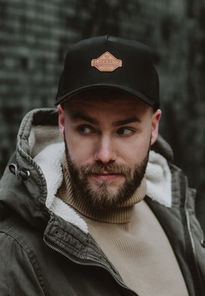 Black trucker cap with a brown leather patch featuring "BLACKSKIES" logo. Worn with a green coat and beige turtleneck.
