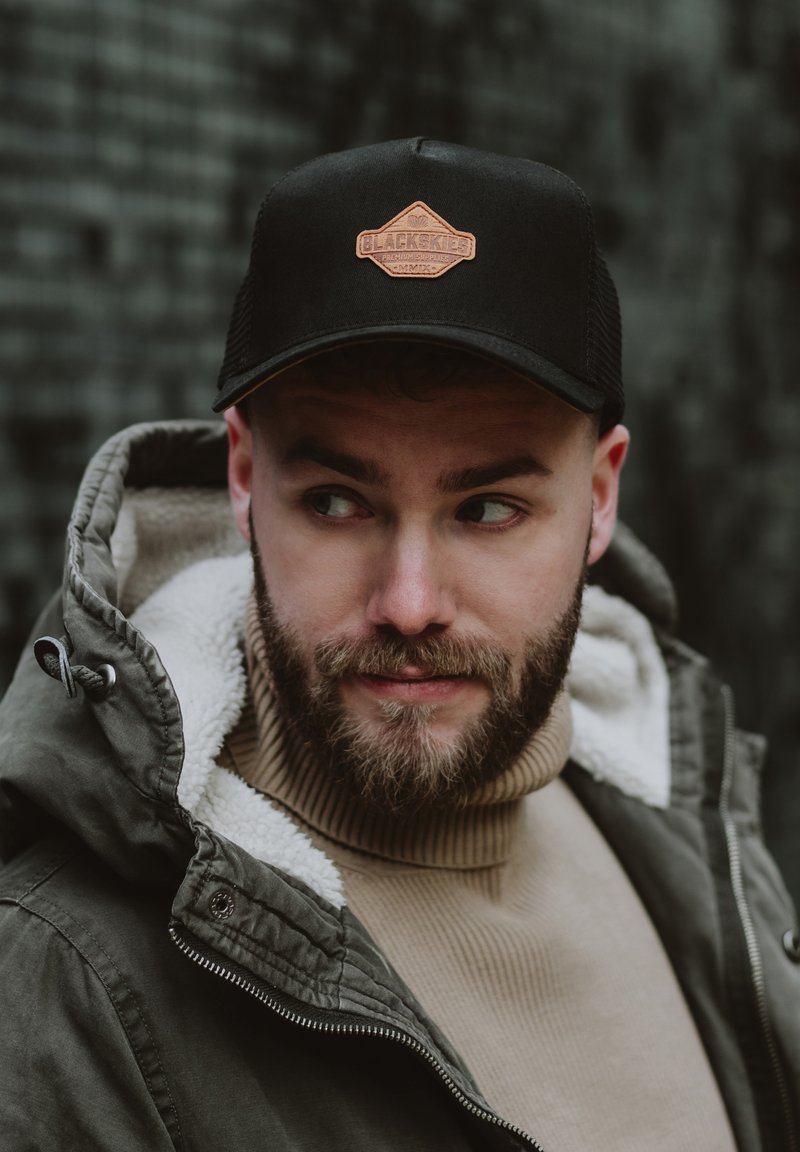 Fekete trucker sapka barna bőrrel ellátott "BLACKSKIES" logóval. Zöld kabáttal és bézs garbóval viselve.