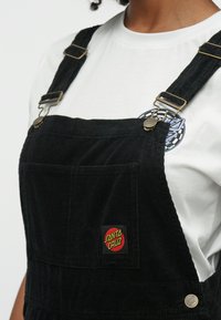 Crne korduroy kombinezone s zlatnim hardverom, s prednjim džepom i crvenom i žutom oznakom "Santa Cruz" na bibliji.