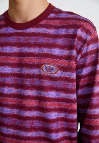 Mörkröd långärmad skjorta med horisontella ränder i lila och blått, med en broderad logotyp i en oval på bröstet. Tillverkad av bomull.