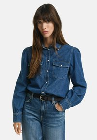 Chemise en denim bleu foncé, avec col à boutons, deux poches à la poitrine et manches longues avec poignets à boutons. Portée avec un jean taille haute.