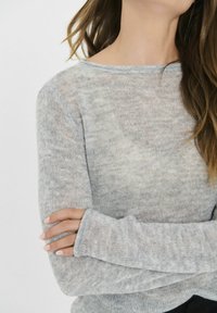Svetlo siva pletena džemper s dugim rukavima, raspuštenom tkaninom, okruglim izrezom i produženim manžetama. Tekstura deluje mekano i blago prozirno.